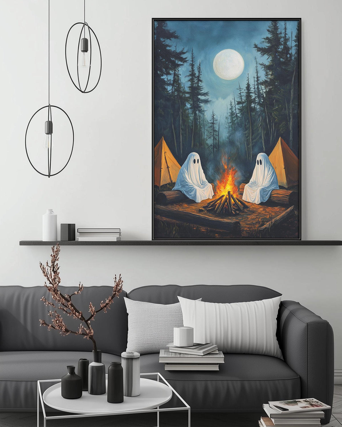 a cozy living room with a large framed painting of a ghostly campfire scene hanging on the wall above a gray couch. The painting depicts two ghostly figures sitting by a campfire, with a full moon illuminating the scene. The room is decorated with various items, including a potted plant, vases, and books, creating a warm and inviting atmosphere.