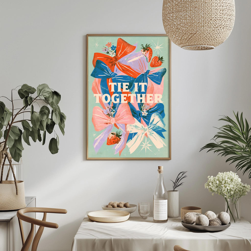 a dining table with a white tablecloth, a bowl of fruit, a bottle, and a vase with flowers. Above the table, there is a framed poster with a colorful floral design and the text "tie it together".