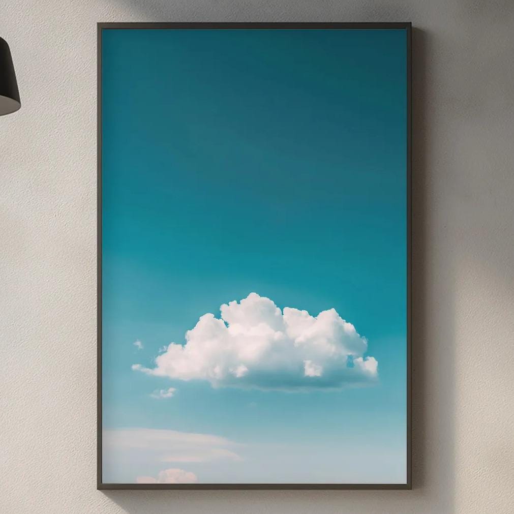 a kitchen counter with various kitchen items, including a cutting board, a soap dispenser, and a plant. Above the counter, there is a framed picture of a blue sky with a fluffy white cloud.