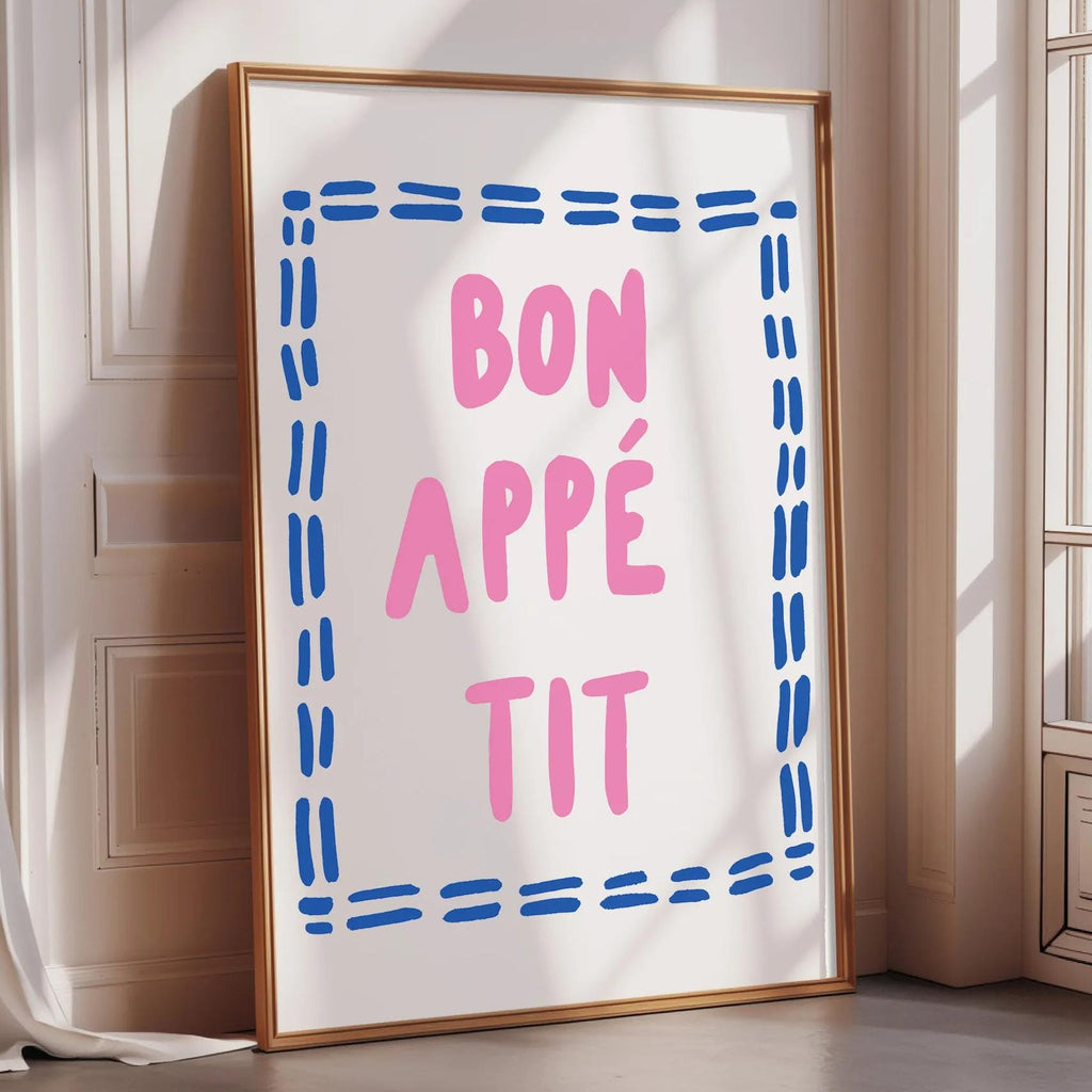 A framed poster with the text " bon appé it" in pink and blue letters, placed on a white wall near a window.