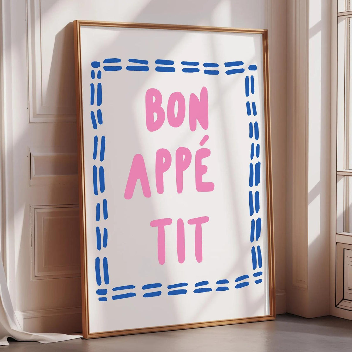 A framed poster with the text " bon appé it" in pink and blue letters, placed on a white wall near a window.