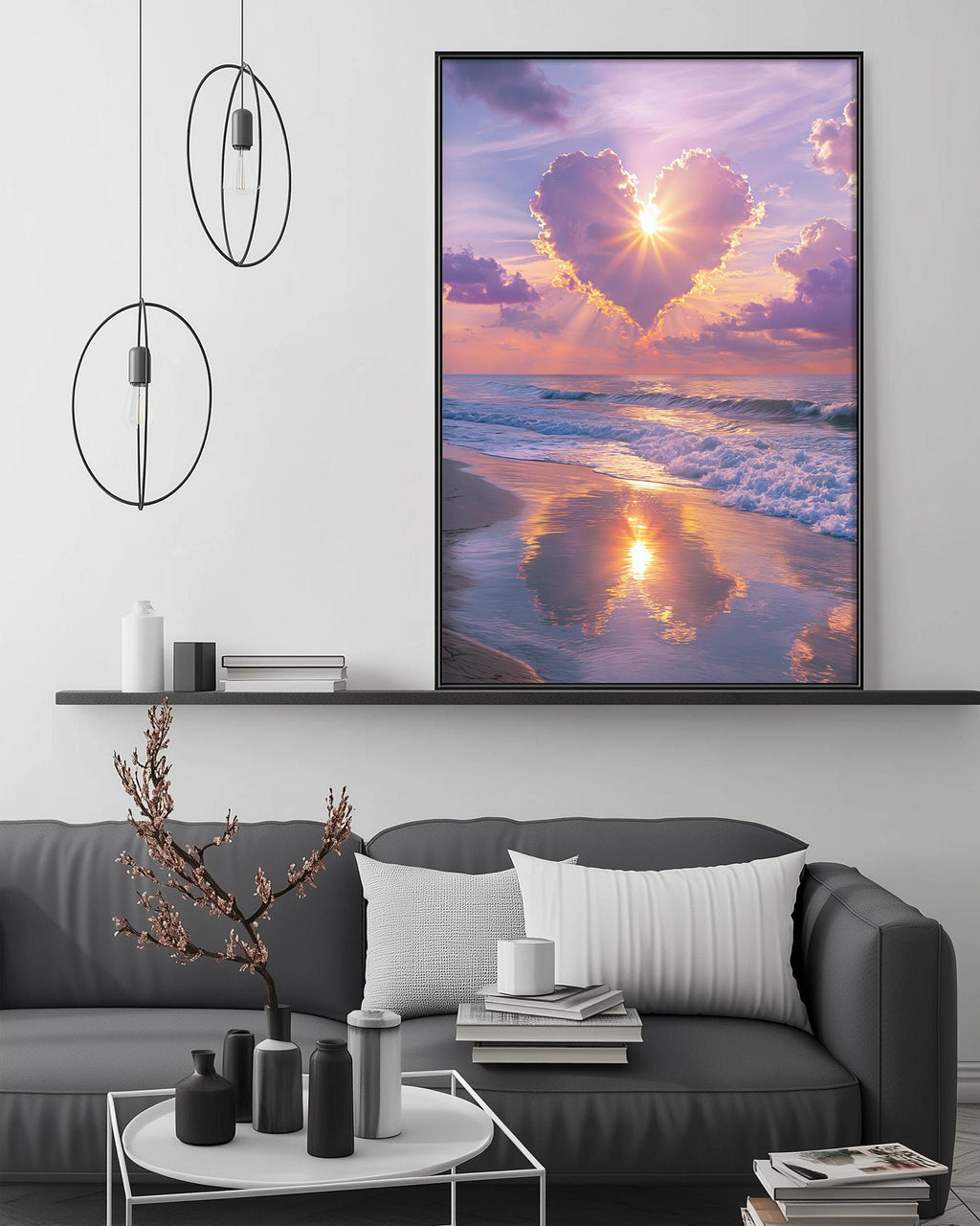 a large framed picture of a serene beach scene with a heart-shaped cloud formation in the sky, reflecting the warm colors of the sunset. The picture is hung on a wall above a gray couch, which has a coffee table in front of it. The room has a minimalist and modern design, with a few decorative items such as a potted plant, vases, and books.