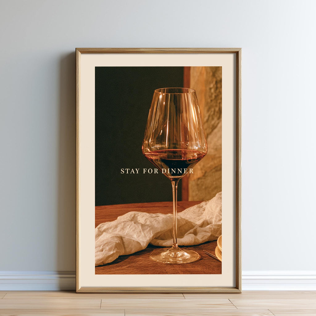 A framed image of a wine glass on a wooden table, with the text "STAY FOR DINNER" displayed on the glass.