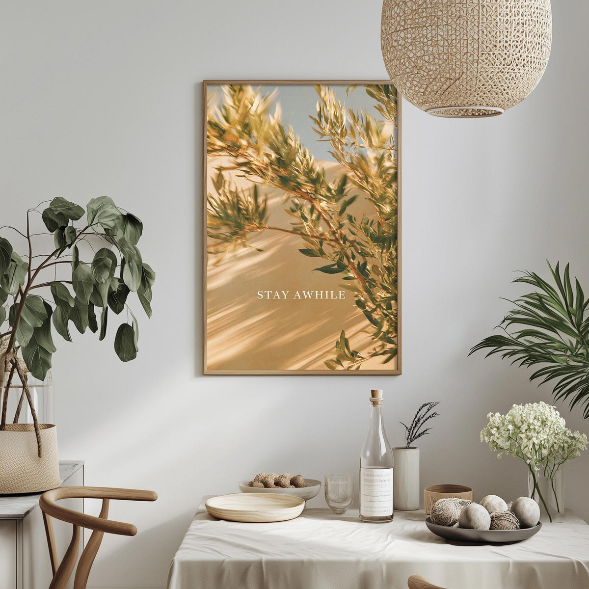 a dining table with a bowl of fruit, a bottle, and a vase of flowers, along with a framed picture hanging on the wall above the table.