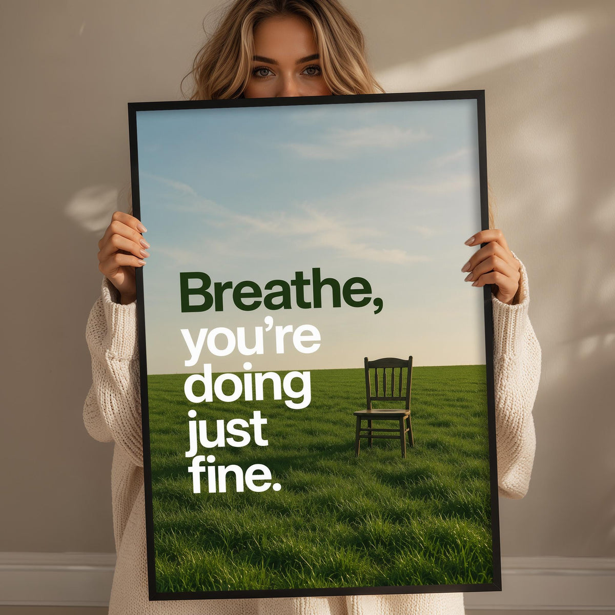 A woman holding a framed poster with a serene image of a chair in a grassy field, along with the text "Breathe, you're doing just fine."