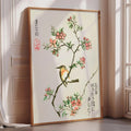 A framed painting depicting a bird perched on a branch with pink flowers, placed on a wooden floor in a room with a window.