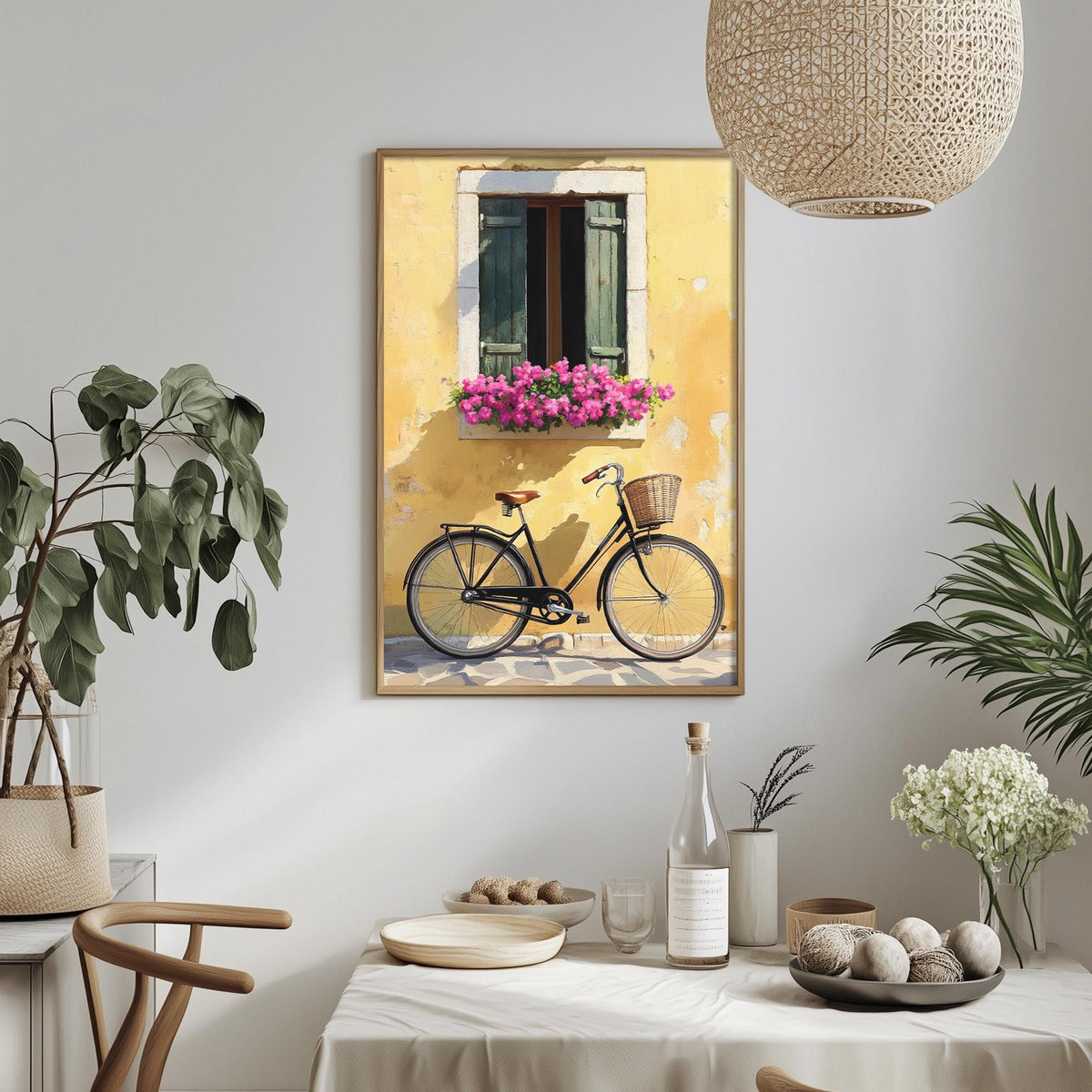 a framed painting of a bicycle parked in front of a yellow wall with a window and flower box, hanging on a wall above a dining table with various decorative items and plants.
