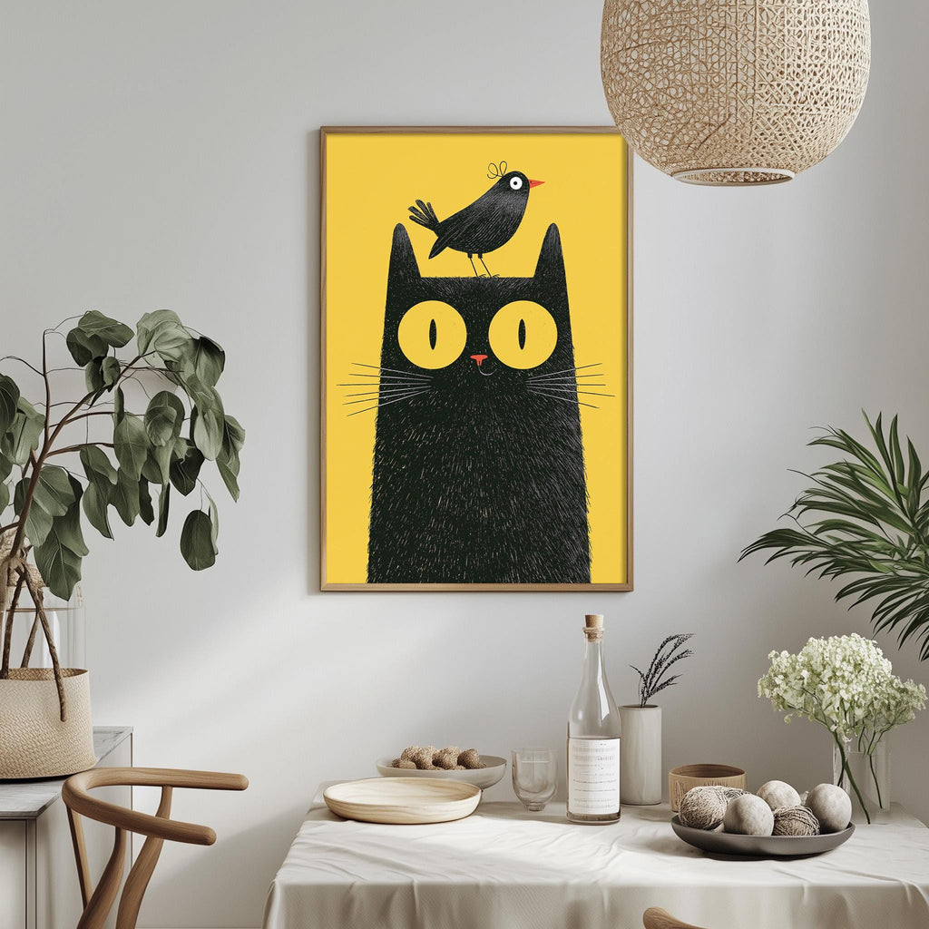 a framed illustration of a black cat with a bird perched on its head, set against a yellow background. The cat and bird are the main subjects of the artwork, which is displayed on a wall above a dining table. The table is set with various items, including a bottle, a bowl, and a vase, creating a cozy and inviting atmosphere.