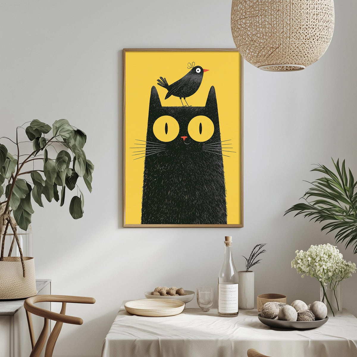 a framed illustration of a black cat with a bird perched on its head, set against a yellow background. The cat and bird are the main subjects of the artwork, which is displayed on a wall above a dining table. The table is set with various items, including a bottle, a bowl, and a vase, creating a cozy and inviting atmosphere.