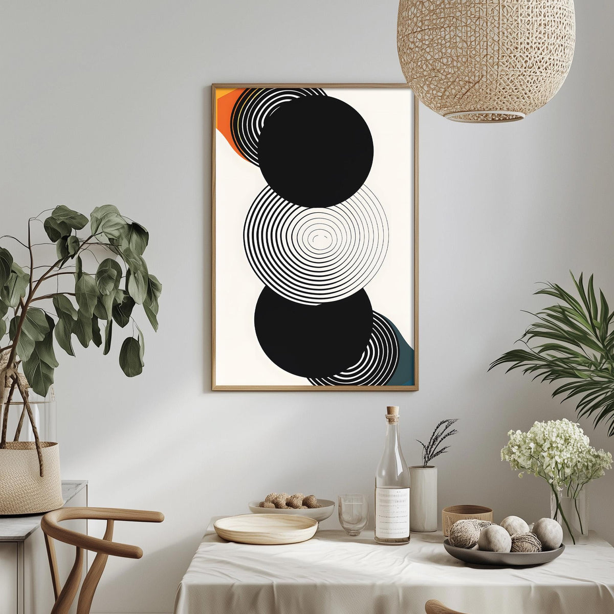 a modern dining room with a minimalist design. The focal point is a framed abstract artwork with black and white circles on a white background. The room is decorated with potted plants, a dining table set with plates and bowls, and a hanging light fixture.