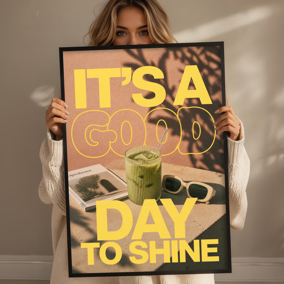 A woman holding a framed poster with the text "IT'S A GOOD DAY TO SHINE" and an image of a green drink, sunglasses, and a magazine.