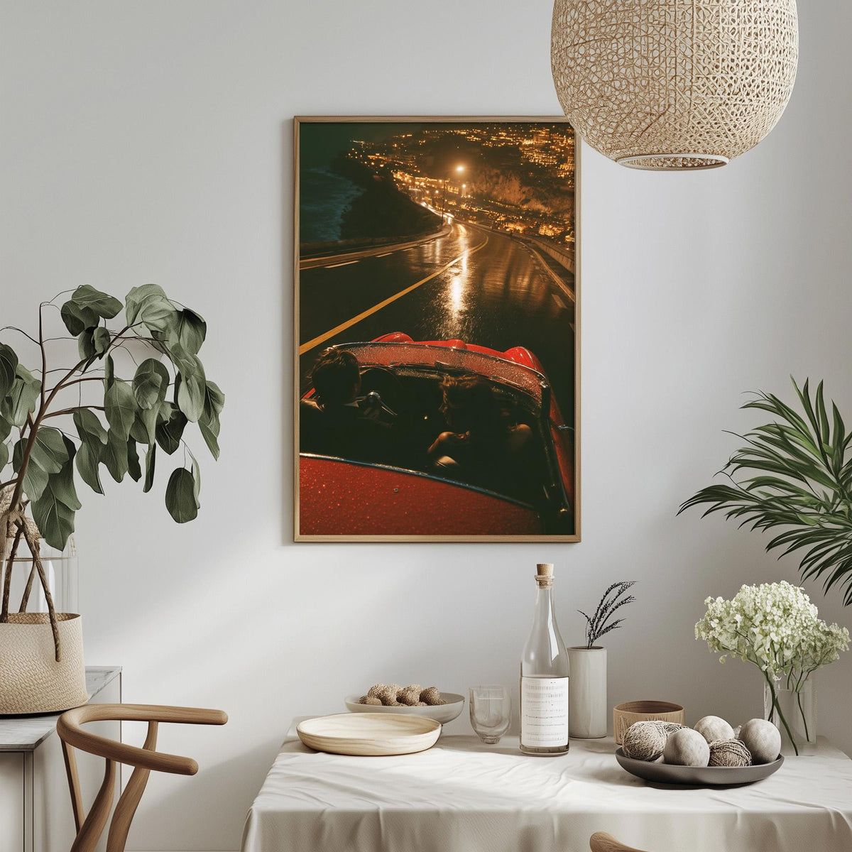 a dining table with a white tablecloth, a bottle of wine, a bowl of fruit, and a vase of flowers. Above the table, there is a framed picture of a red car on a road at night, with a city skyline visible in the background.