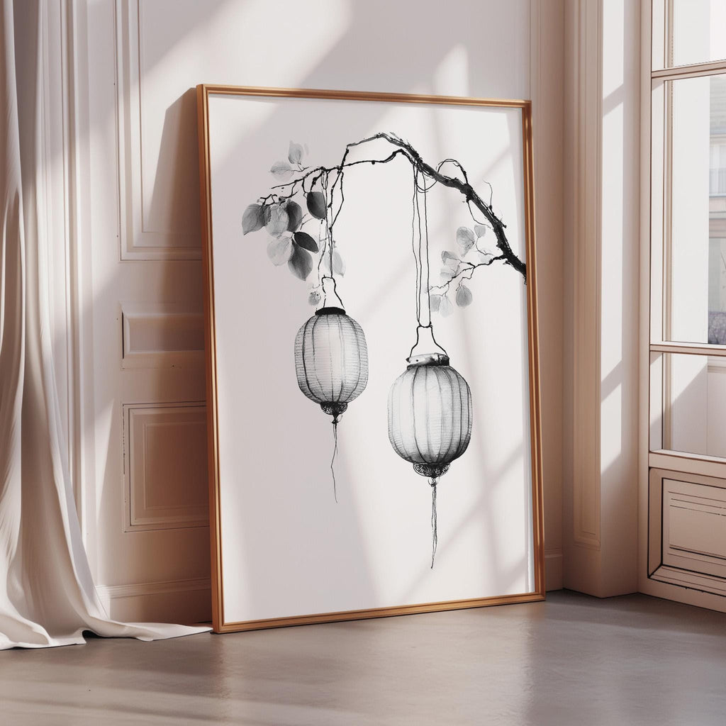 A framed black and white illustration of two lanterns hanging from a branch, placed on a wooden floor in a room with a window.