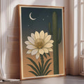 a framed illustration of a cactus with a large white flower on its side, set against a night sky with a crescent moon and stars.