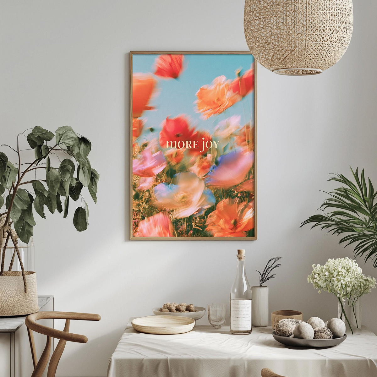 a dining table with a white tablecloth, a bowl of fruit, a bottle, and a vase with flowers. Above the table, there is a framed poster with a vibrant floral design and the text "MORE JOY".