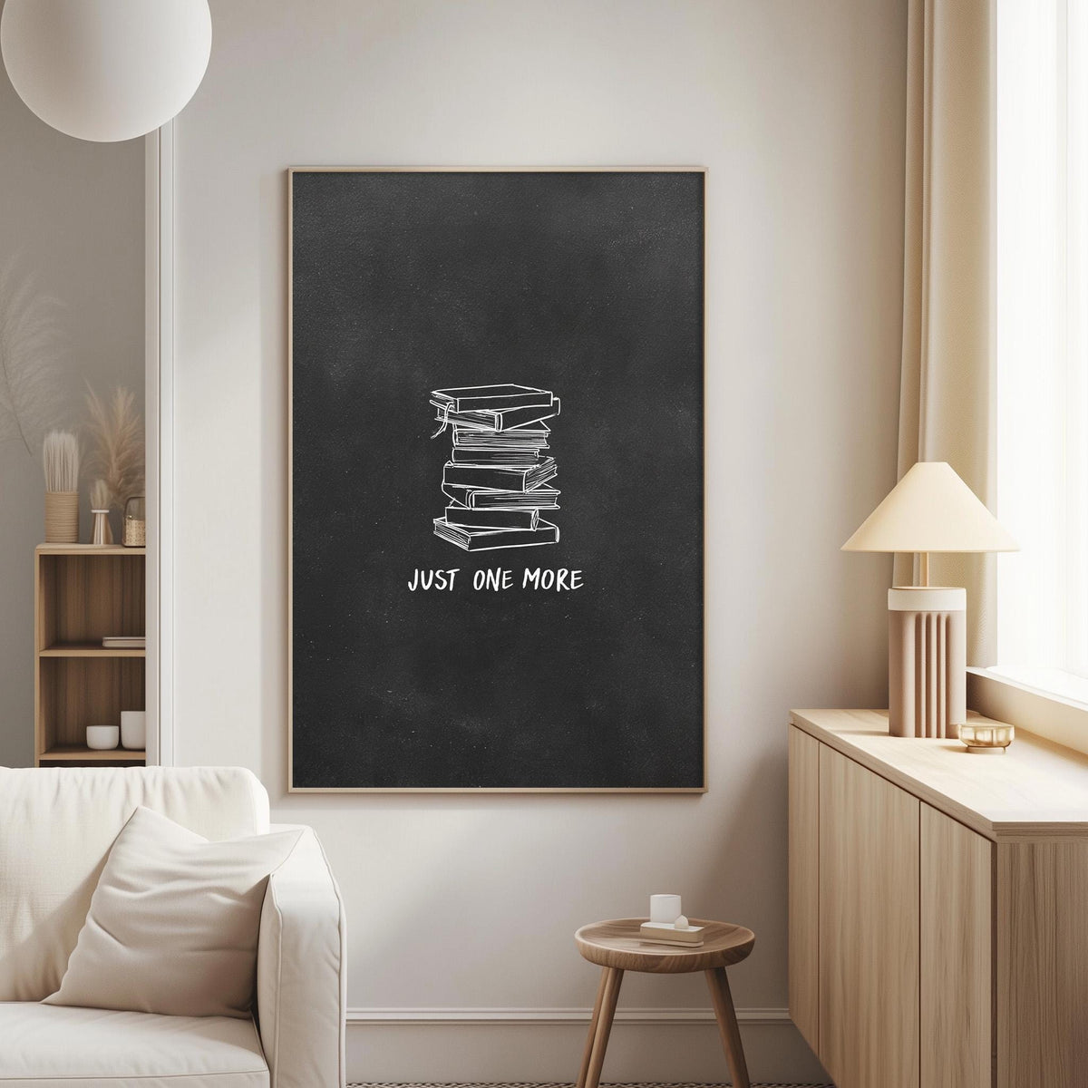 a living room with a white couch, a wooden side table, and a lamp. Above the couch, there is a blackboard with a white chalk drawing of a stack of books and the text "JUST ONE MORE" written on it.