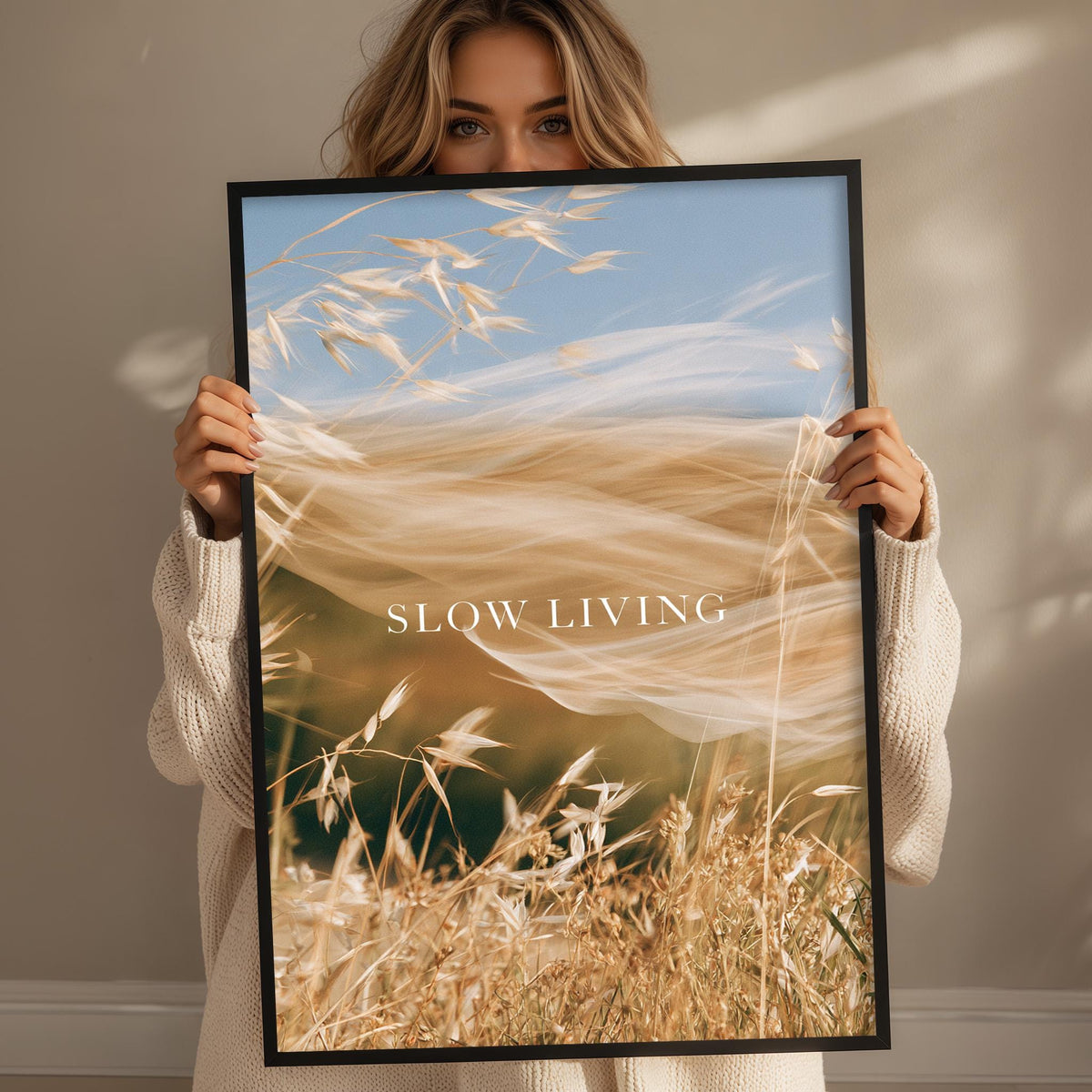 A woman holding a framed print with a blurred image of a field of tall grass and the text "SLOW LIVING" on it.