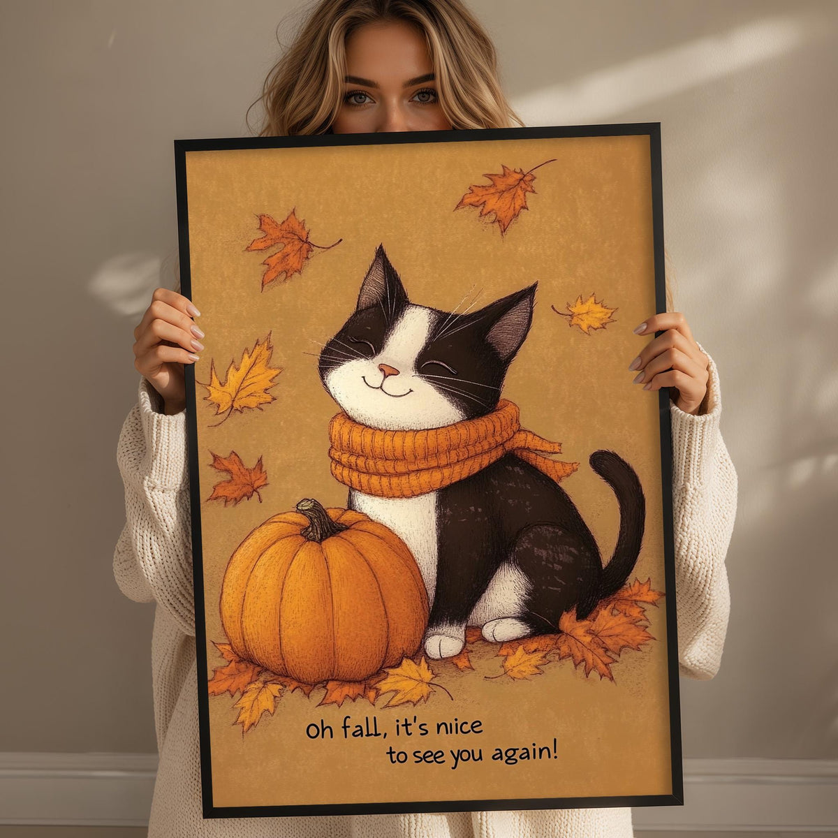A woman holds up a framed illustration of a black and white cat sitting next to a pumpkin, surrounded by autumn leaves.