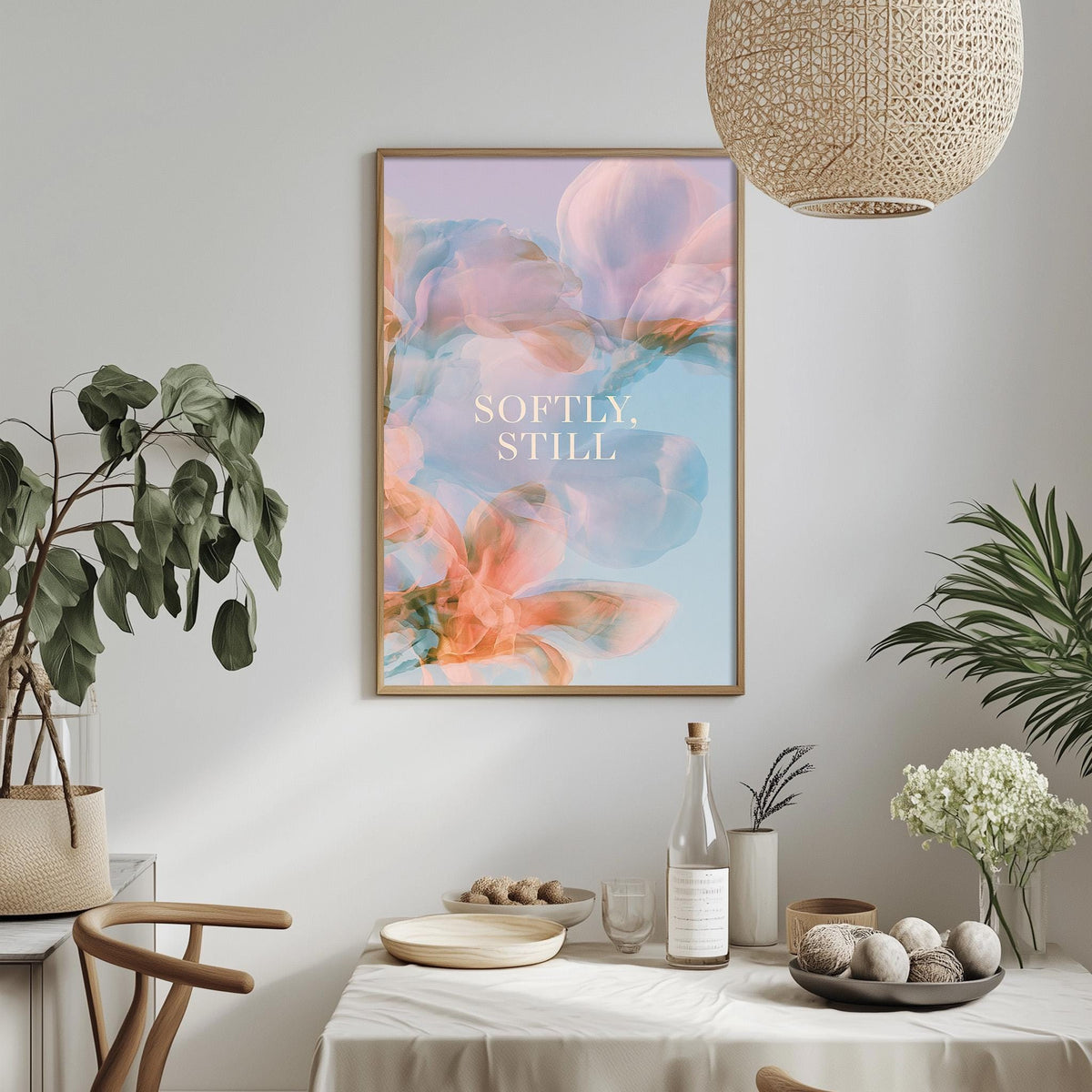 a dining table with a white tablecloth, a bowl of fruit, a bottle, and a vase with flowers. Above the table, there is a framed poster with the text "Softly Still" in the center.
