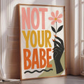 A framed poster with the text "NOT YOUR BABE" and an illustration of a hand holding a flower.