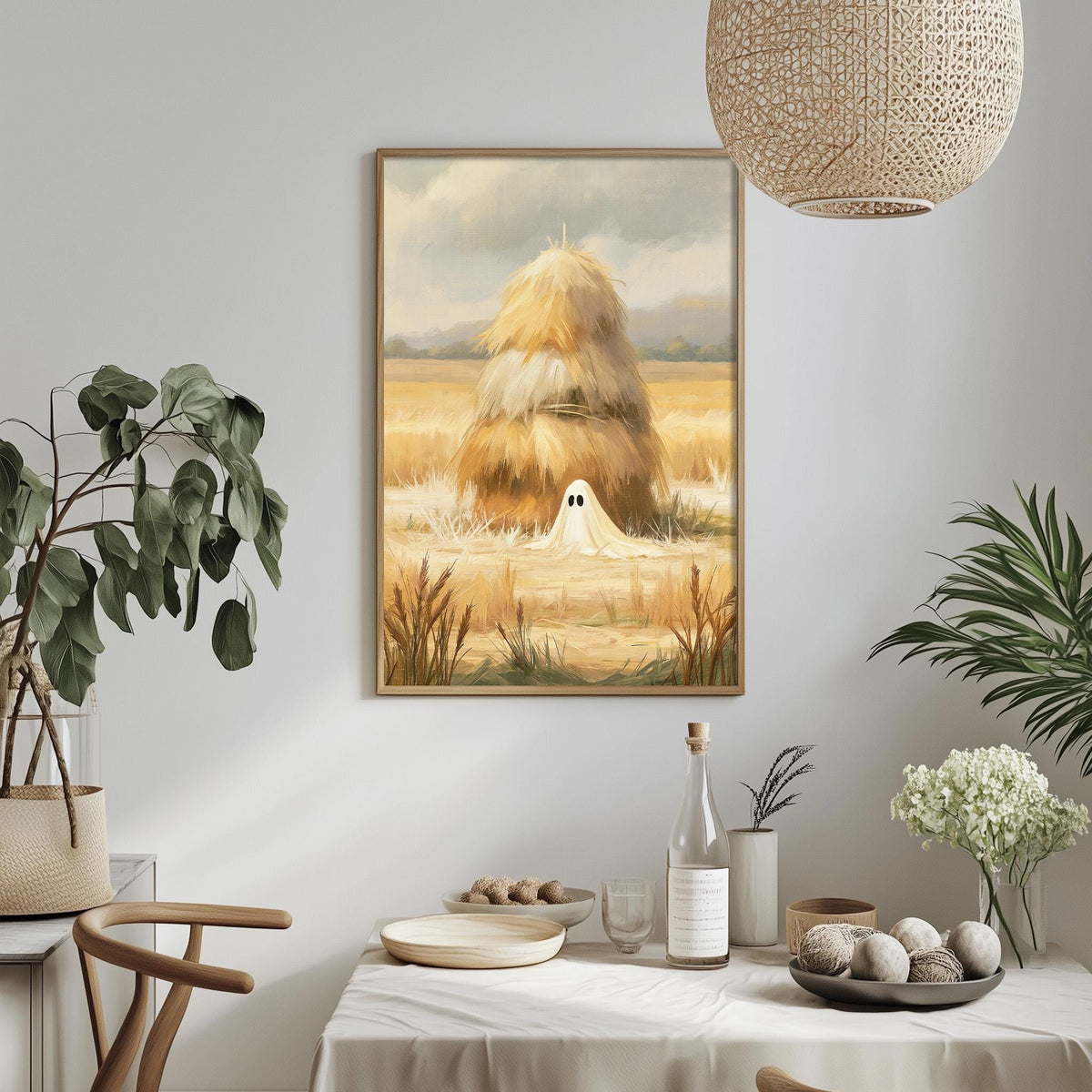 a dining table with a white tablecloth, a bowl of fruit, a bottle, and a vase with flowers. Above the table, there is a framed painting depicting a ghost in a field with hay bales.