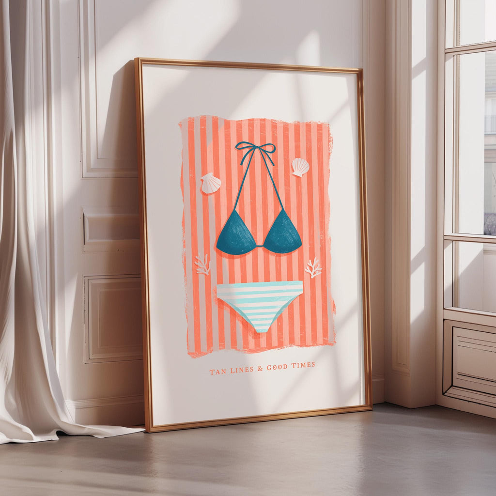 A framed poster with a beach scene, featuring a bikini top, a striped towel, and seashells, along with the text "TAN LINES & GOOD TIMES".