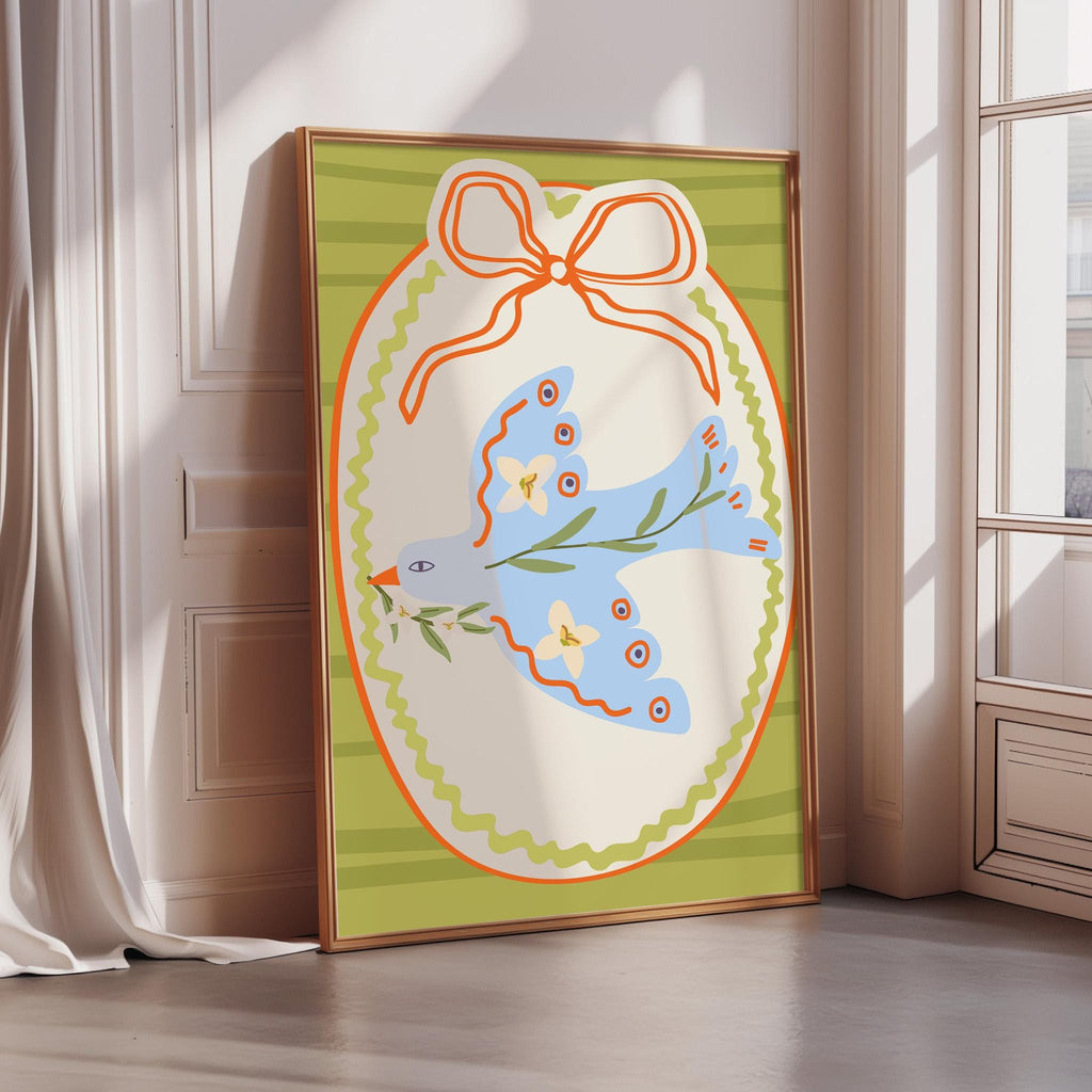 A framed illustration of a blue bird with a bow on a green and white background, placed on a wooden floor next to a window.
