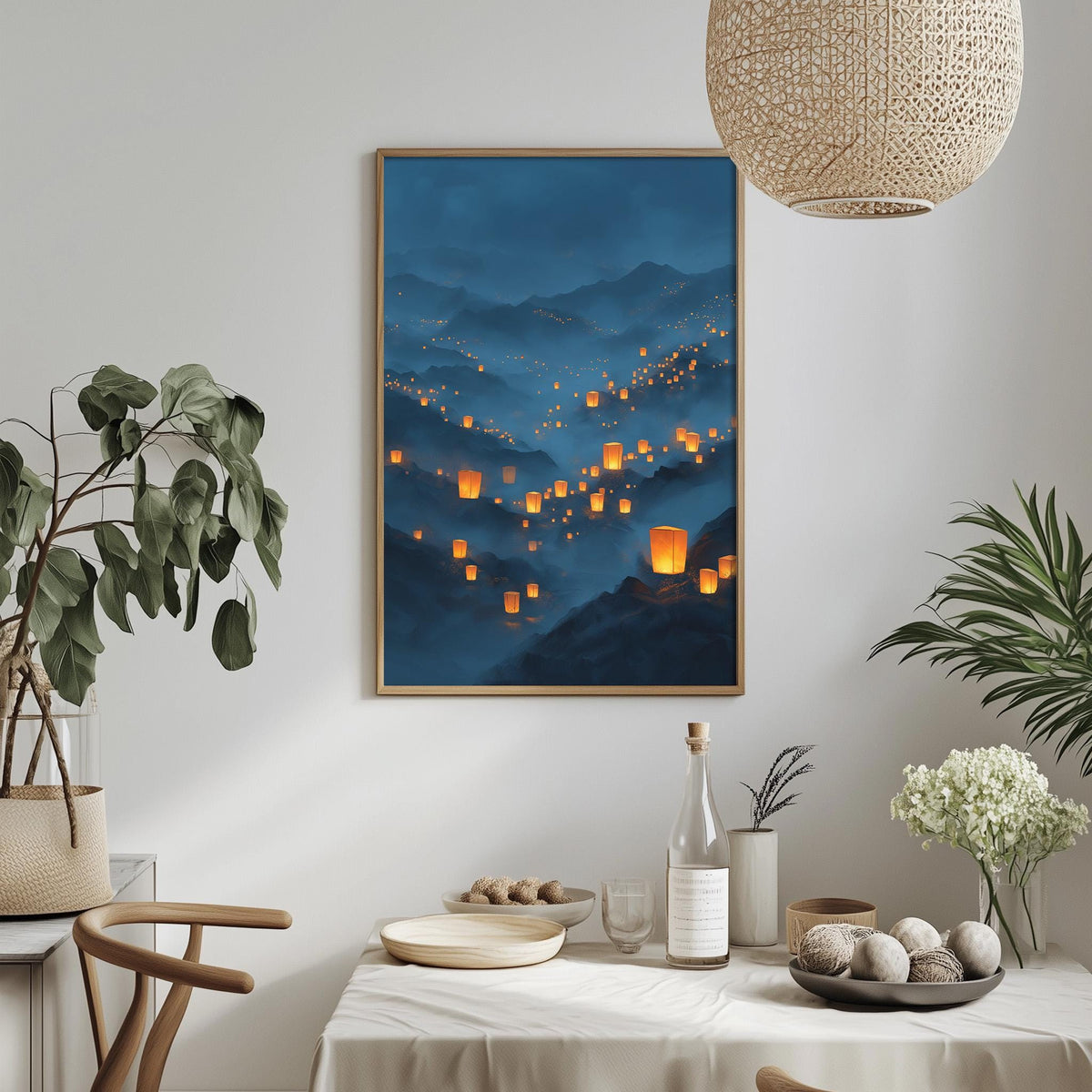 a dining table with a white tablecloth, a bottle, a bowl, and a vase with flowers. Above the table, there is a framed picture of a landscape with orange lanterns floating in the sky.