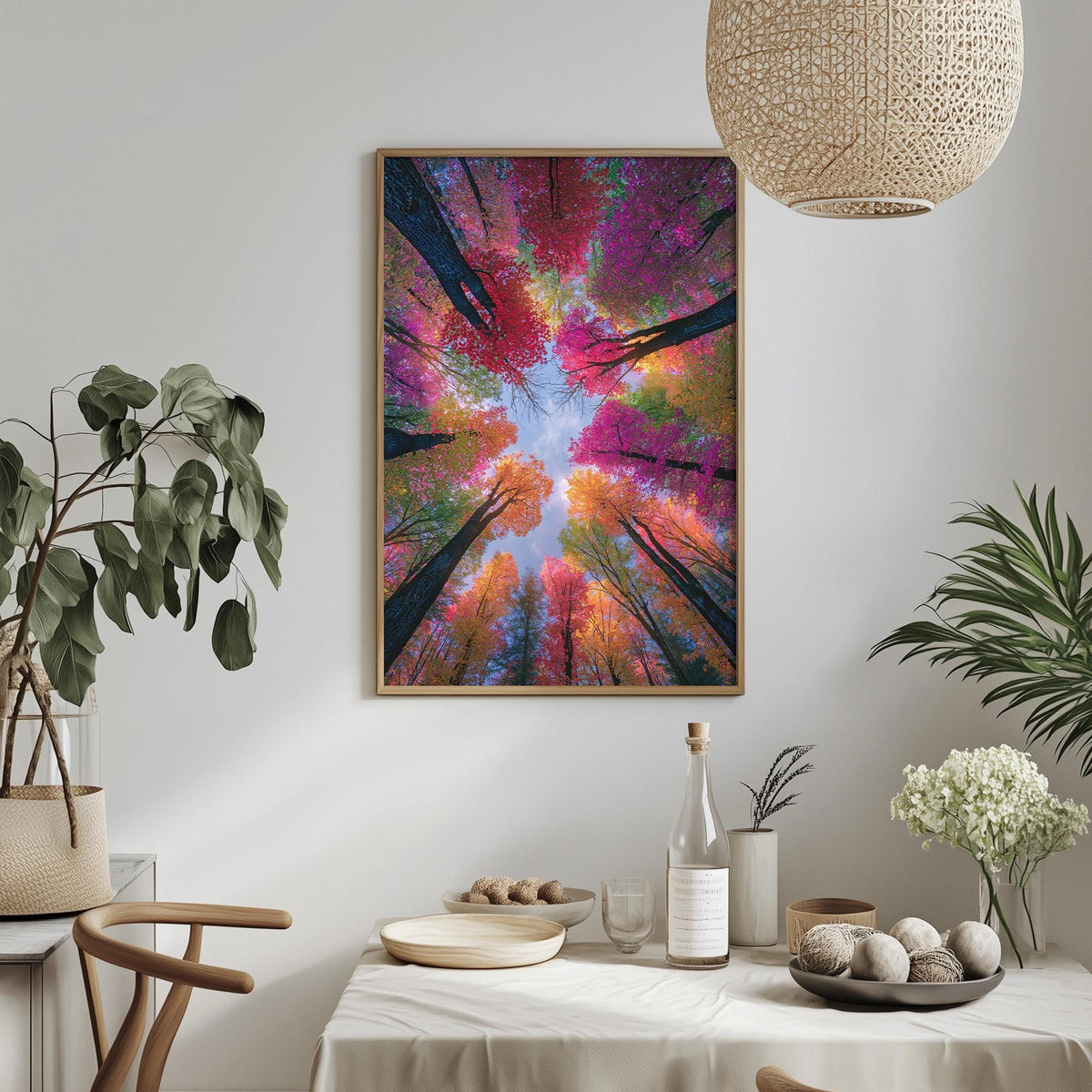 a dining table with a white tablecloth, a bowl of fruit, a bottle, and a vase with flowers. Above the table, there is a framed picture of a forest with a vibrant, colorful sky.