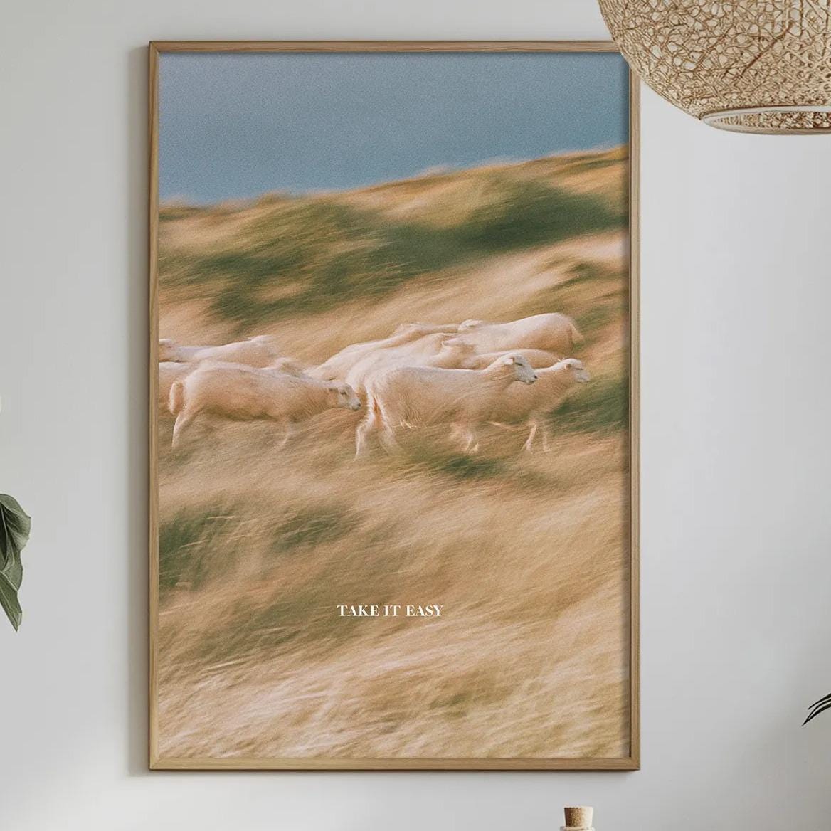 a dining table with a white tablecloth, a bowl of fruit, a bottle, and a vase with flowers. Above the table, there is a framed picture of a field with sheep.