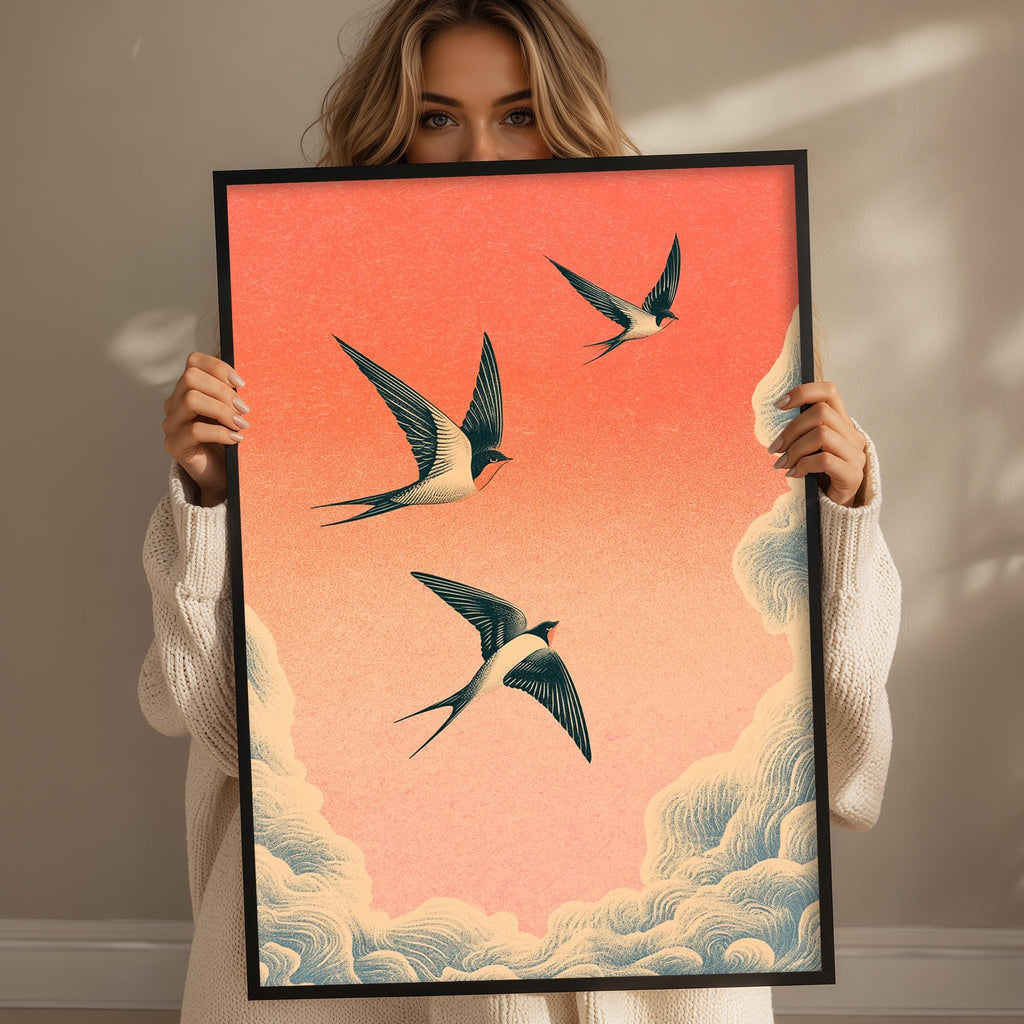 A woman holds up a framed print of three birds in flight against a vibrant orange and pink sky.