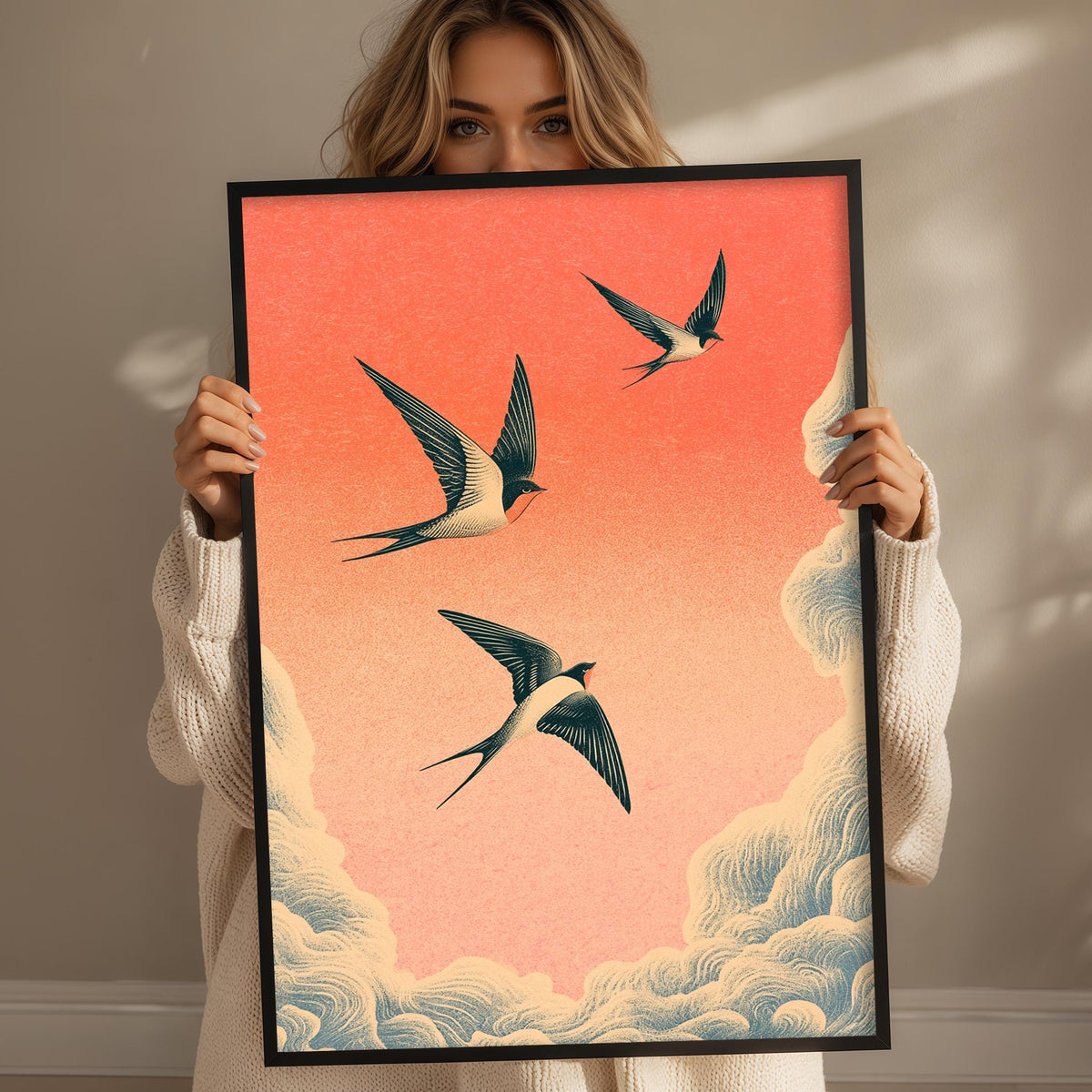 A woman holds up a framed print of three birds in flight against a vibrant orange and pink sky.