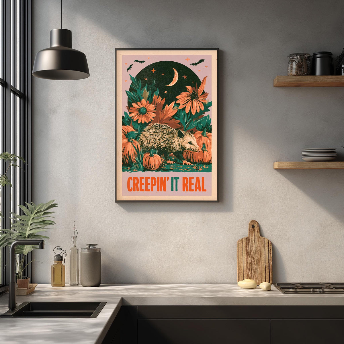 a framed poster with a whimsical illustration of a hedgehog surrounded by flowers and pumpkins, with the text "CREEPIN' IT REAL" displayed prominently. The poster is hanging on a wall above a kitchen counter with various kitchen items such as a cutting board, a bottle, and a bowl.