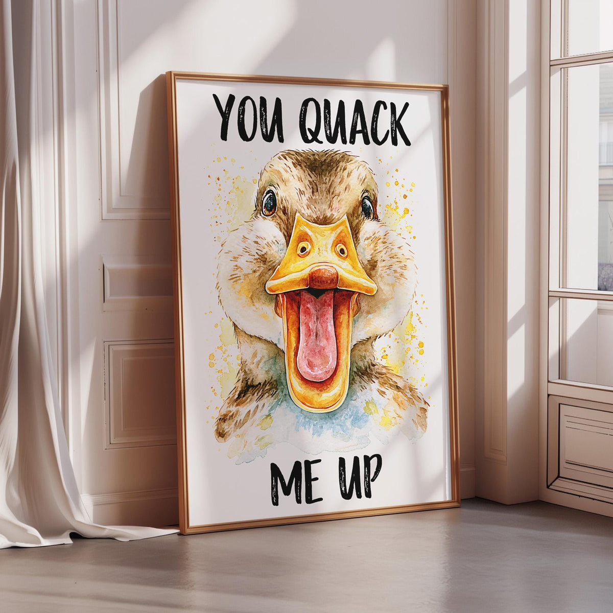 A framed poster with a duck's face and the text "YOU QUIACK ME UP" on it, placed on a wooden floor next to a window.