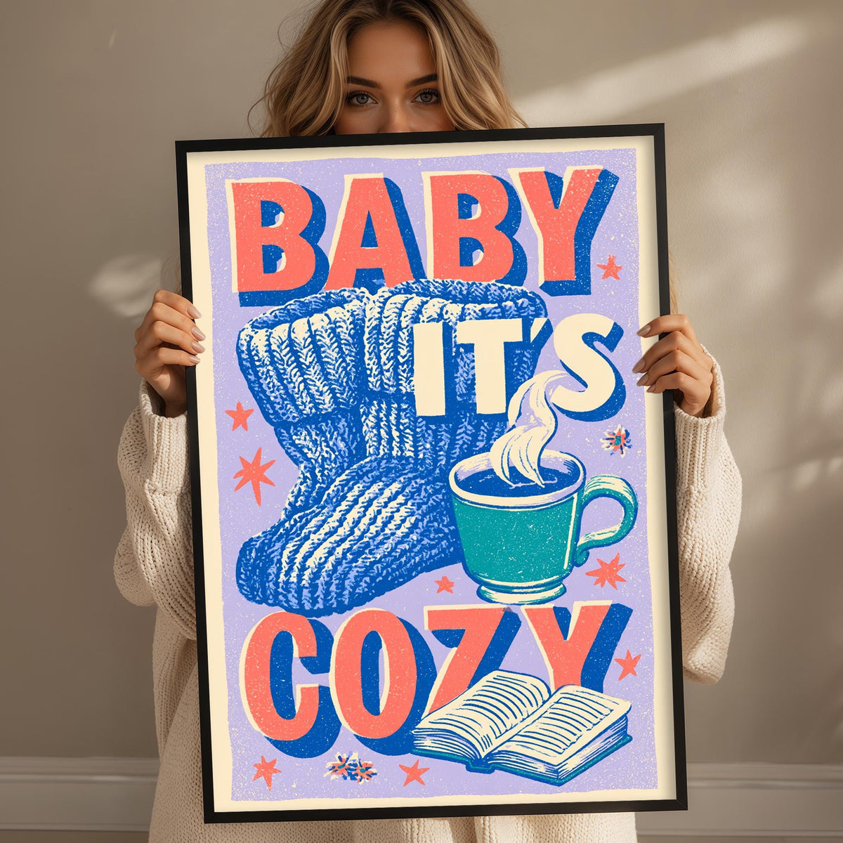 A woman holding a framed poster with the text "BABY IT'S COZY" and an illustration of a mug with a hot beverage.