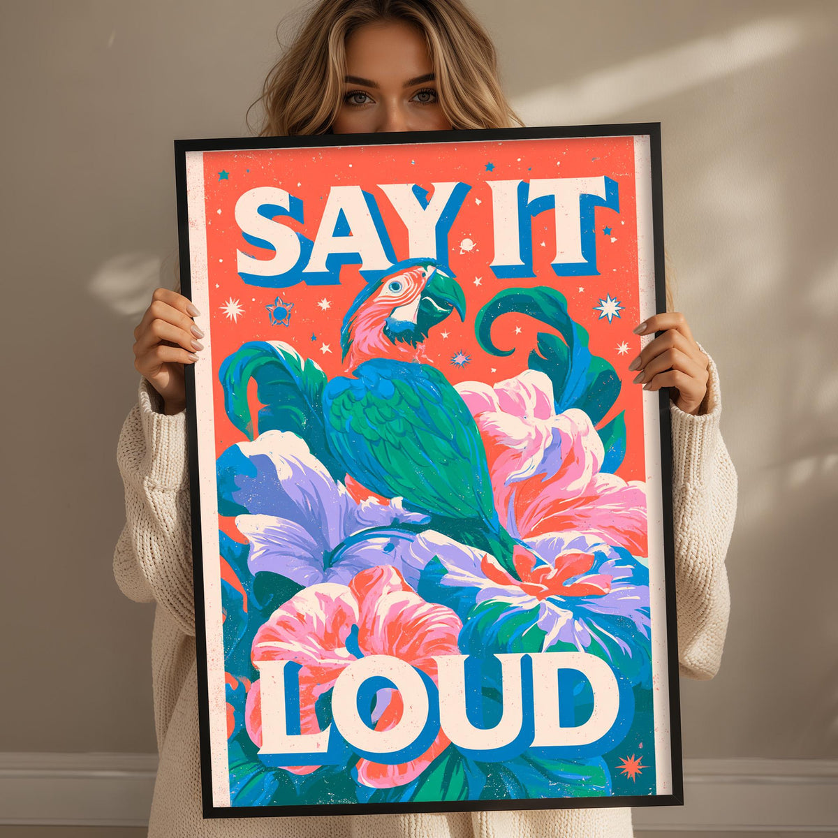 A woman is holding up a framed poster with a colorful illustration of a parrot and flowers. The poster has the text "SAY IT LOUD" written on it.