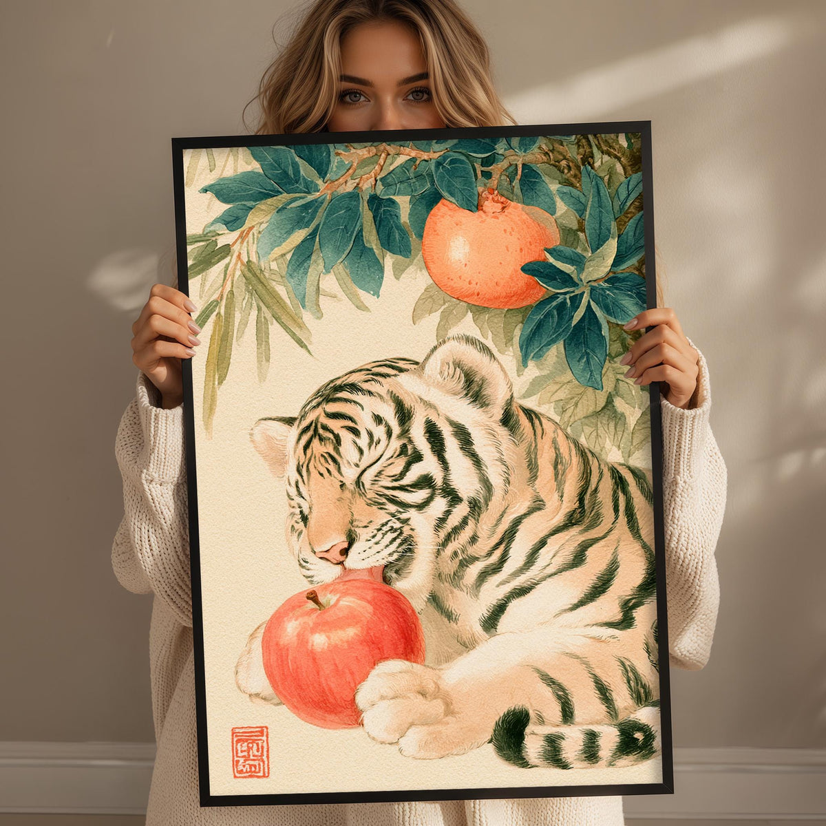 A woman holds up a framed painting of a tiger resting on an apple tree, with the tiger's head resting on an apple.