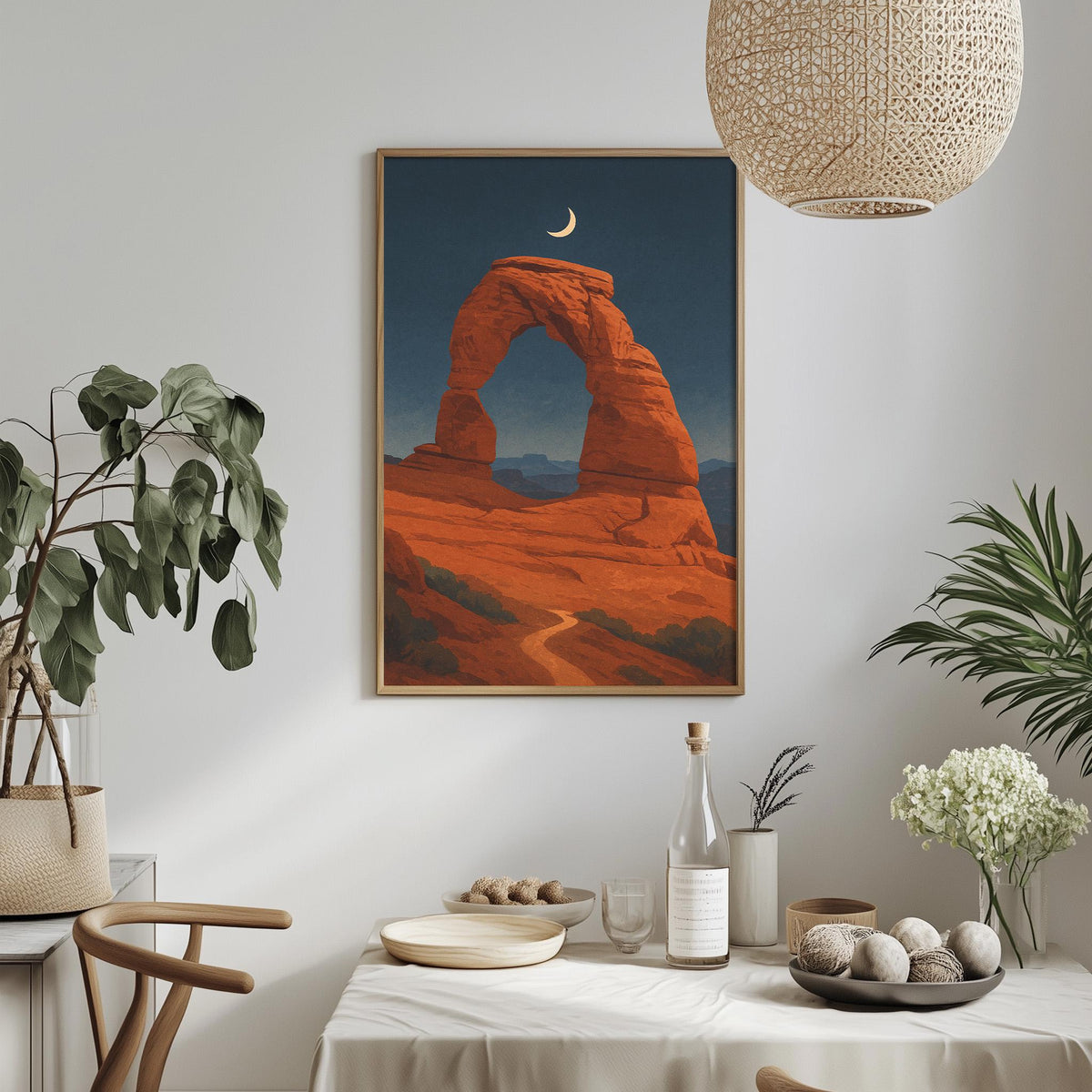 a framed painting of a desert landscape with a crescent moon in the sky, hanging on a wall above a dining table with various decorative items and plants.