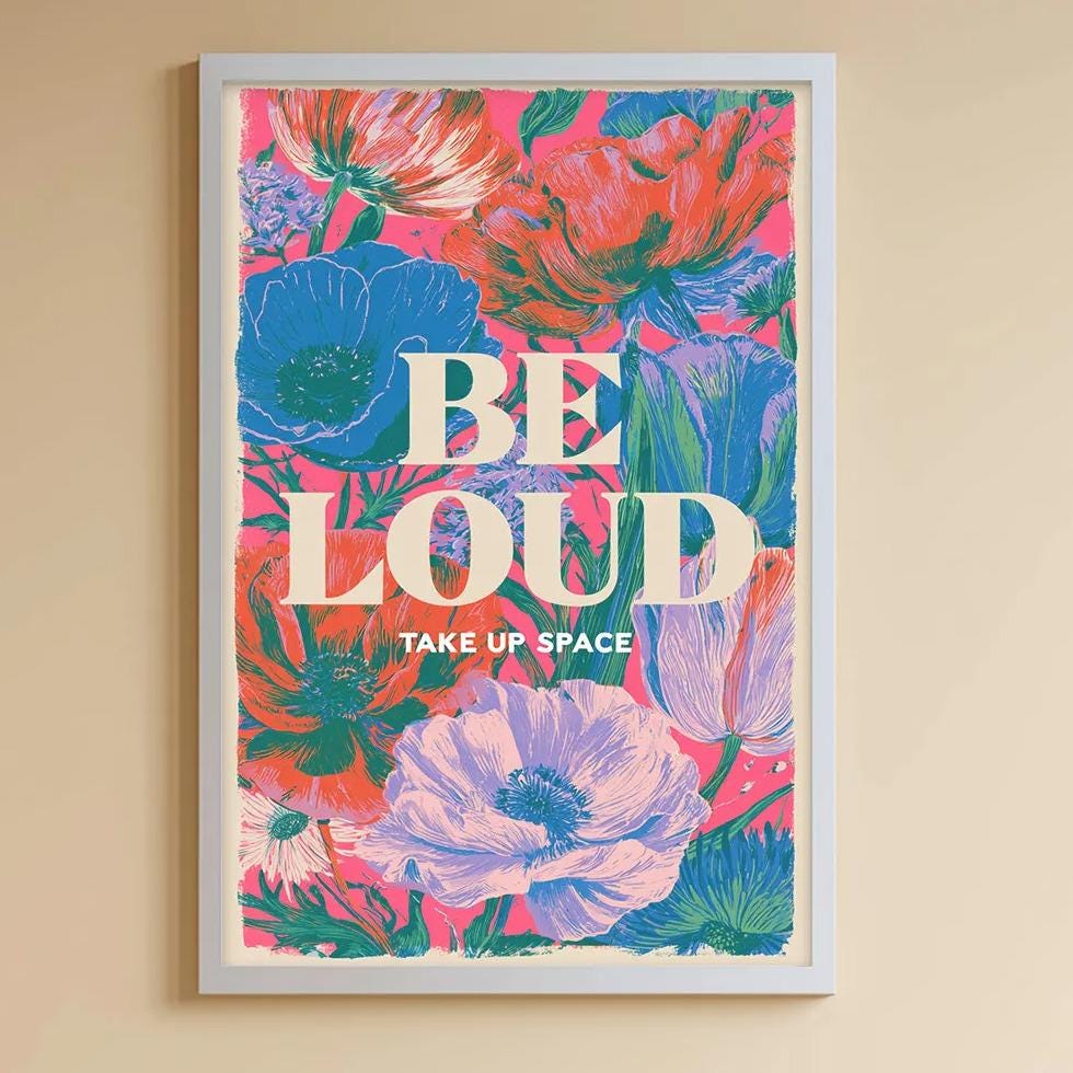 A child's bedroom with a white bookshelf, a stuffed rabbit, and a colorful poster on the wall. The poster has the text "BE LOUD TAKE UP SPACE" and features a floral design.