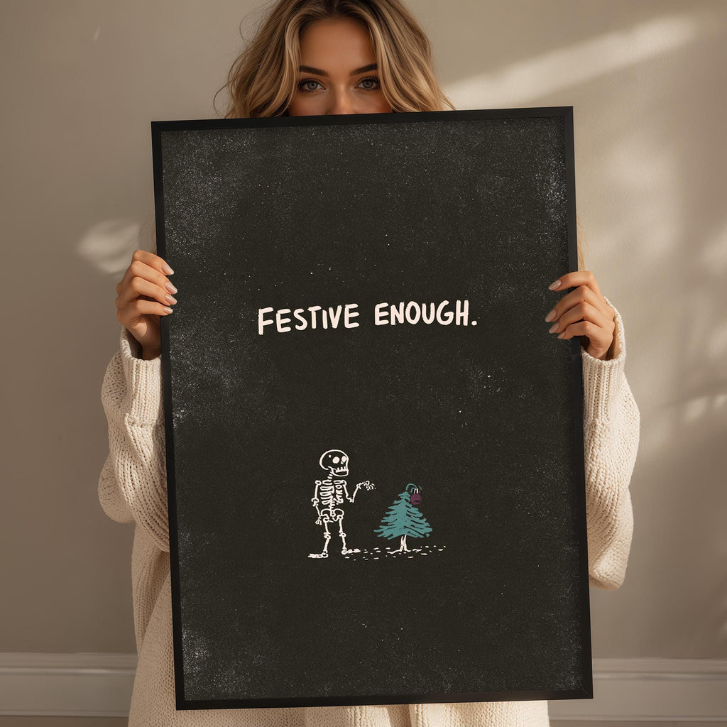 A woman holding a blackboard with the text "FESTIVE ENOUGH" written on it. Below the text, there is a drawing of a skeleton and a Christmas tree.