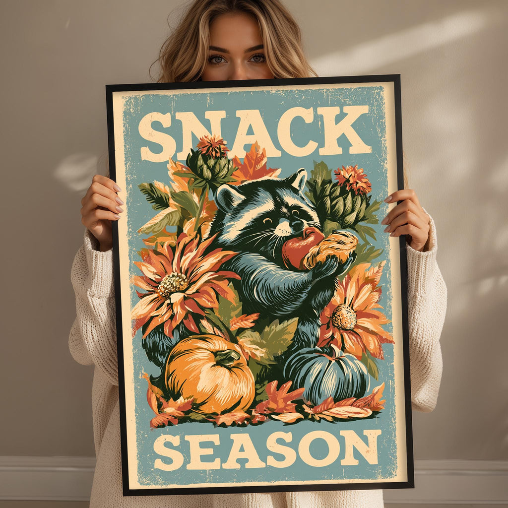 A woman is holding up a framed poster with a raccoon and various autumn-themed elements, including flowers and pumpkins.