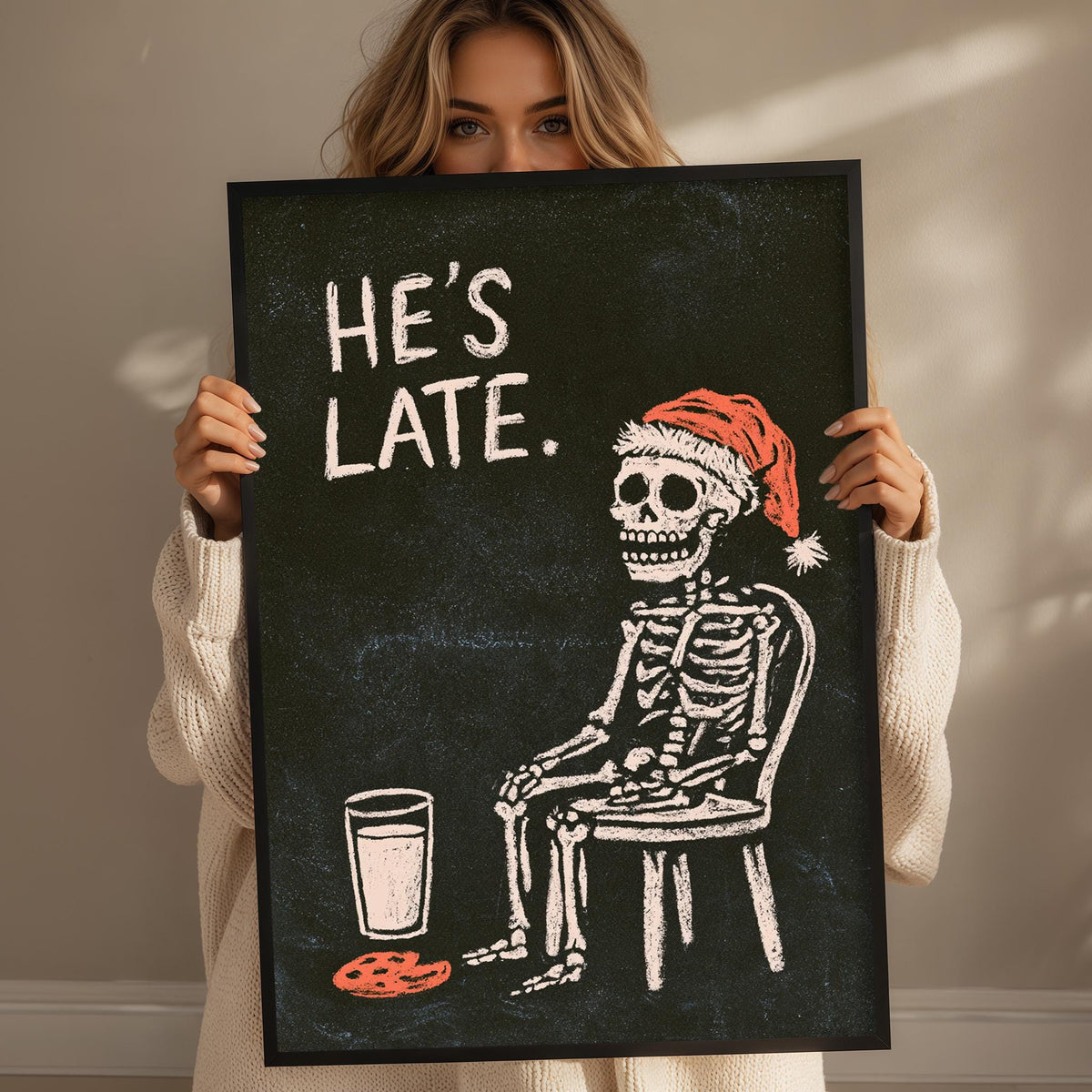 A woman is holding up a framed image that depicts a skeleton wearing a Santa hat and sitting in a chair, with a glass of milk next to it.
