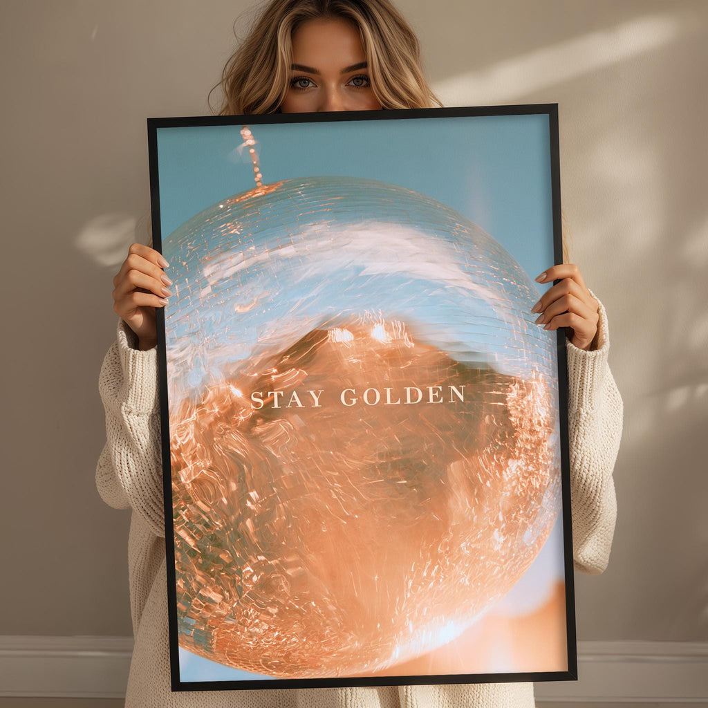 A woman holding a framed poster with a large, colorful image of a globe and the text "STAY GOLDEN" on it.