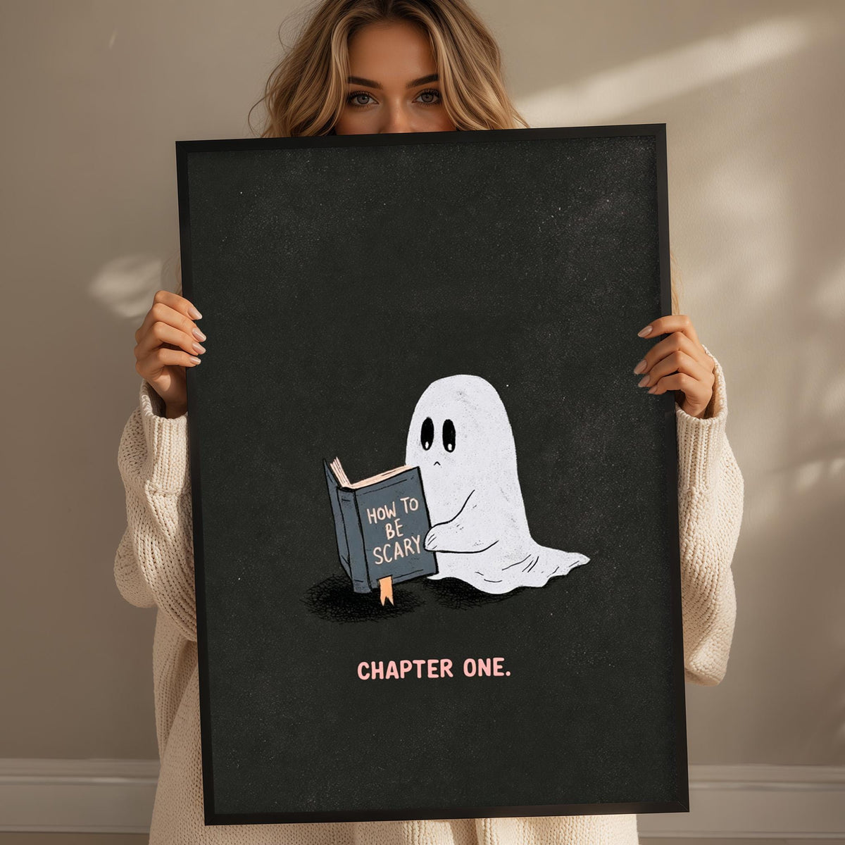 A woman holding a framed poster with a ghost reading a book titled "How to be scary" and the caption "CHAPTER ONE."