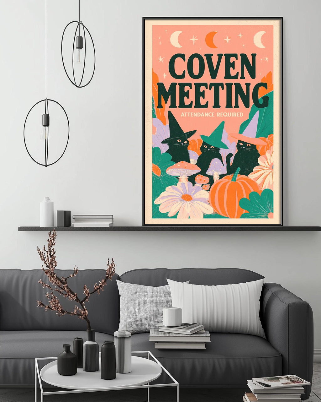 a living room with a gray couch, a coffee table, and a framed poster on the wall. The poster has a whimsical design with cats, flowers, and a crescent moon, and it reads "COVEN MEETING ATTENDANCE REQUIRED".