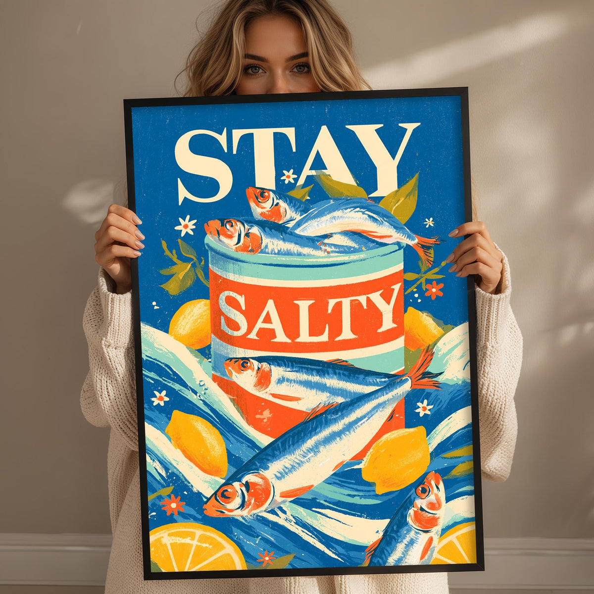 A woman is holding up a framed poster with a vibrant design featuring fish, lemons, and the text "STAY SALTY".