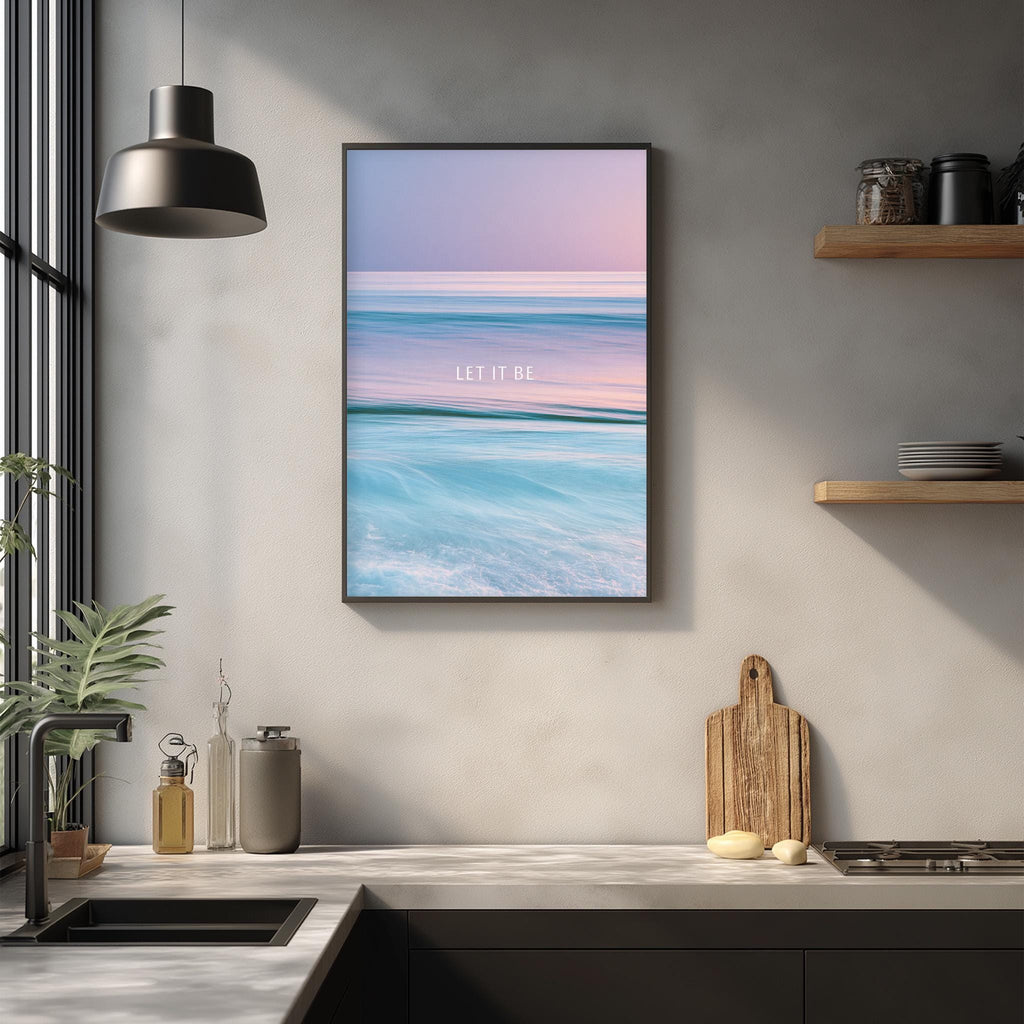 a kitchen counter with a framed poster of a beach scene hanging above it. The poster has a serene beach landscape with waves and a sunset. The kitchen counter has various items on it, including a cutting board, a bottle, and a vase. The overall scene is calming and peaceful, reflecting the beach setting depicted in the poster.