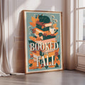 A framed poster with a cat sitting on top of a stack of books, surrounded by autumn leaves, flowers, and a cup of coffee. The text on the poster reads "BOOKED FOR FALL".