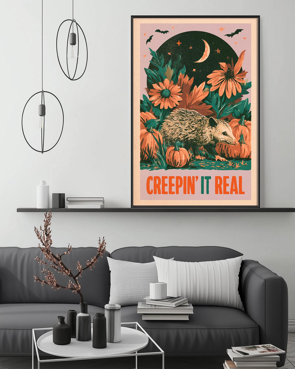 a living room with a gray couch, a coffee table, and a framed poster of an opossum surrounded by flowers and pumpkins. The poster has a whimsical and spooky design, with the text "CREEPIN' IT REAL" prominently displayed.