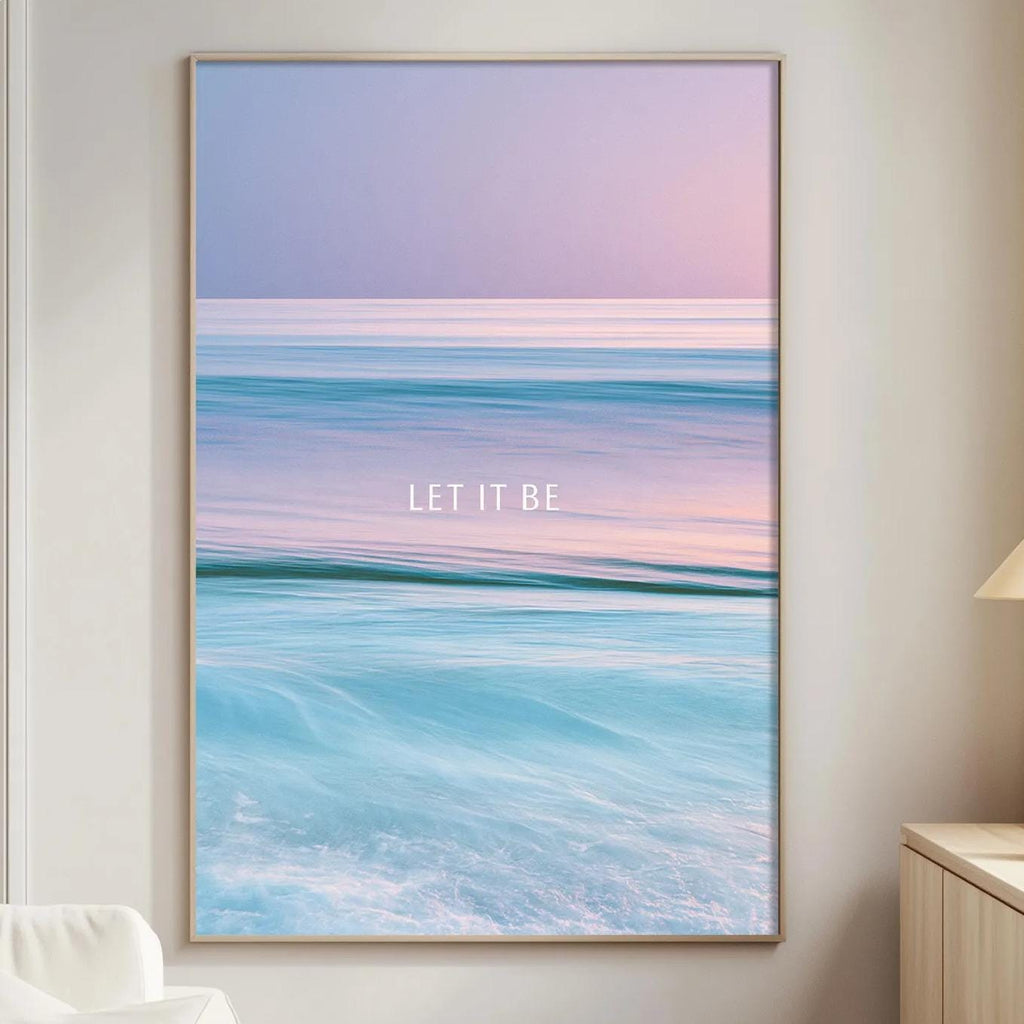 a large framed poster of a seascape with the text "LET IT BE" displayed prominently on it. The poster is hanging on a wall in a living room setting, with a white couch and a small table nearby.